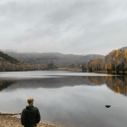 Man standing on shoreline looking out at the calm river surrounded by trees and mountains